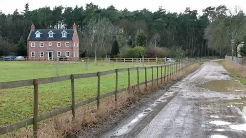 Evelyn Simak/Geograph A view of the former hamlet of West Harling. There is a rough track on the right, with deep puddles. On the left is a wooden fence and a paddock. On the left is a four storey brick house and behind it all are mostly evergreen trees.