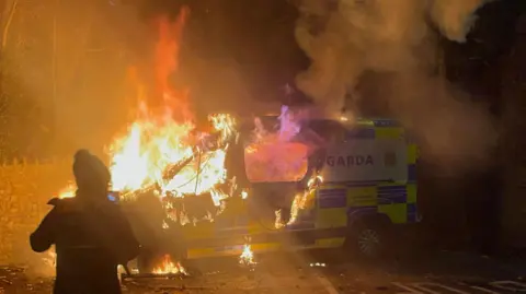 Getty Images A burning police vehicle at night. There is a person stood in front of the vehicle. The vehicle has 'GARDA' written on it.  