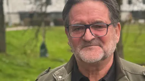 Mike Marshall, a man standing on the campus at the University of East Anglia, The background is slightly blurred and he is standing in the foreground. He is looking directly at the camera and is wearing a black shirt with a khaki jacket over the top and a pair of glasses over his eyes.