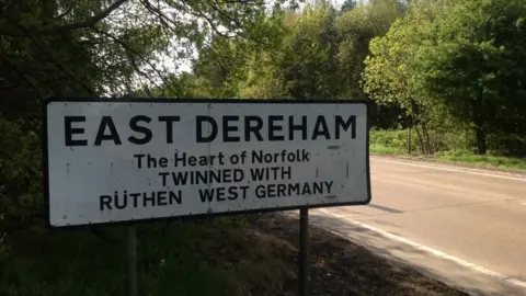 The roadsign by the A47 at Dereham, bearing the words: "East Dereham. The heart of Norfolk. Twinned with Ruthen, West Germany. It is on the side of the single carriageway section, with trees either side of the road.