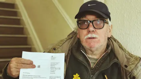 LDRS Arthur Bowling is wearing a black baseball cap, glasses, a green top, two thicker tops and a coat, while holding a letter from the Department for Work and Pensions about his application for pension credit.