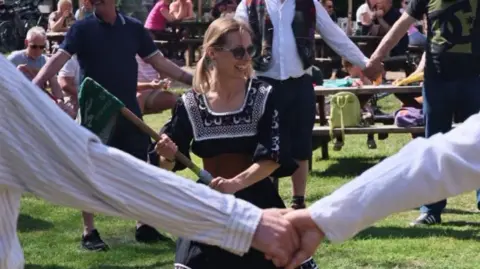 A woman stands in the middle of a circle of people. She is holding a large stick in both hands which has a green rag attached to the end. She is smiling. She wears sunglasses and a black dress. People watching on from the other side of the circle. 