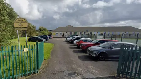 View of the entrance to Bransty Primary School. Cars are parked either side of an entrance road, with grassed area and shrubs on the left. The school, a low-rise building with a pitched roof can be seen in the background under a cloudy sky.