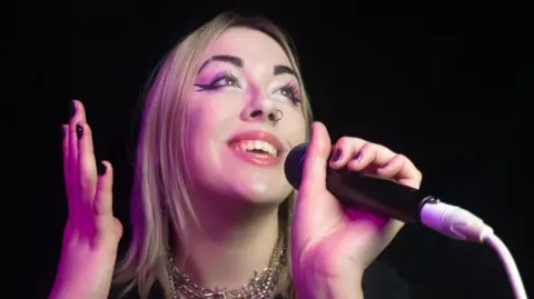 Jade Maris Jade has silver-blond hair and thick black eyeliner. She has a silver nose ring and black nail polish on, she is singing into a mic close up to the camera.