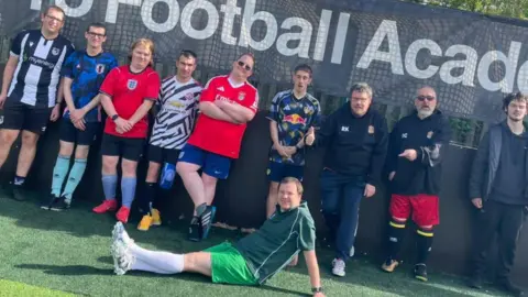 Sheffield Mencap and Gateway A group of men wearing various different football shirts and shorts are stood in a row on a playing field. There is netting saying Football Academy in the background