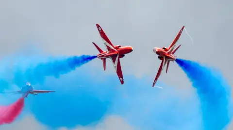 The Red Arrows perform at RAF Fairford during the Royal Military Air Tattoo on July 15, 2023 in Fairford. Two red aircraft trailing blue smoke perform a close pass with a third aircraft with red smoke in the background.