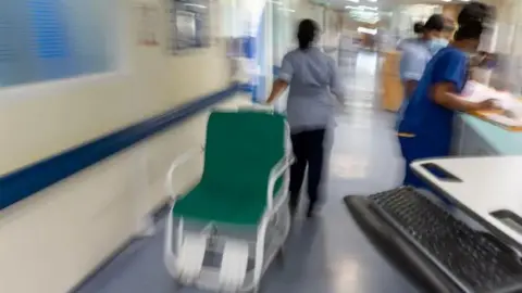 PA Media An image that is out of focus showing medical staff working in a hospital ward, with one nurse pulling an empty green wheelchair while others do paper work at a bench.