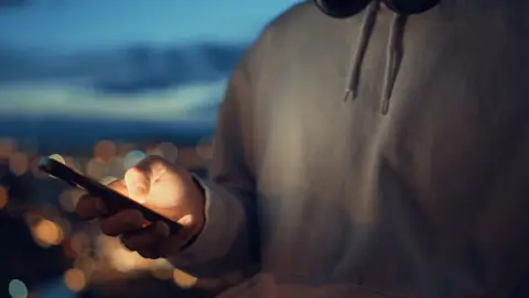 A young person in a grey hoodie can be seen on their phone at night time. 
Their face is not visible and they are wearing headphones. 