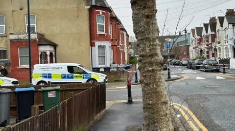 A police van with "Forensic Services" on the side parked on a road within a police cordon.