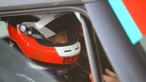Supplied Close-up view of a person sitting inside a race car, wearing a bright red, black, and white full-face helmet with a clear visor. The helmet has visible branding and small printed text. The person is secured in a racing seat with harness straps, and part of the car’s interior and window frame is visible. Another hand appears in the foreground near the door frame, possibly assisting or adjusting something. Lighting is soft with reflections on the helmet visor and car surfaces.
