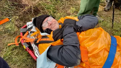 Professor Chris Lavy is wrapped up in an orange blanket on a stretcher and is smiling after breaking his ankle