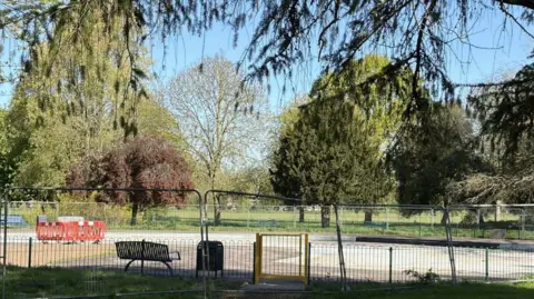 Richard Madden/BBC A concrete paddling pool area behind a temporary metal fence. Trees, barriers and a large grassed area are also visible.