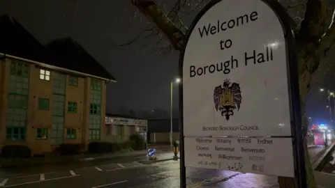Martin Heath/BBC The outside of Bedford Borough Council's offices at night, with a large sign saying "Welcome to Borough Hall", with an eagle crest below and "welcome" in several languages. There is a road running past, with clear signs of rainwater, and a three-storey brick-built office building opposite.