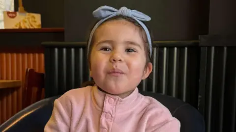 Louisa Palmisano smiles wearing a pink button-up jumper with a blue headband. She is sat in a chair next to a radiator indoors. 