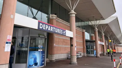 Exterior view of an airport terminal departure entrance, featuring tall pink stone columns, a glass-fronted facade and a large canopy roof. A prominent blue “DEPARTURES” sign hangs above the entrance doors, while a shuttle bus, airport staff and roadside bollards are visible along the curbside drop-off area.