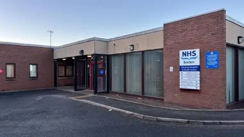 A brick hospital building with an NHS Borders sign on the front and a tarmac path and pavement in front of it