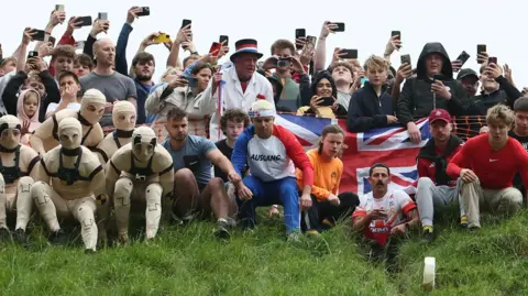 Reuters Crowds of people are standing at the top of a hill, some with their phones out filming what is happening below, and others bracing themselves to run down the hill after a wheel of cheese. Some people are dressed up, others are wearing T-shirts and jogging bottoms.