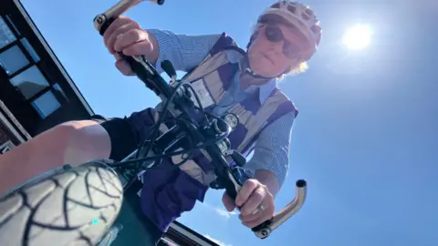 Jon Wright/BBC A woman on a bike, the photo is taken from the view of the front wheel. the sun is blazing in a blue sky. She is wearing a cycle helmet and sunglasses.