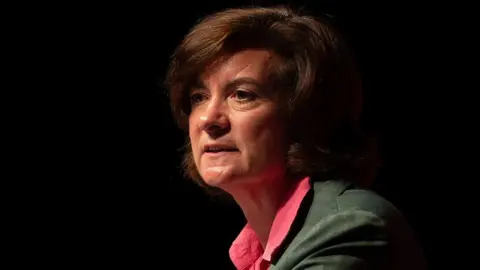 A side-shot of Eluned Morgan in front of a dark background, wearing a green suit and a red shirt. She is looking out into space in front of her.
