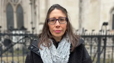 BBC Kristie Higgs, pictured outside the Court of Appeal. She has shoulder-length brown hair, and wears dark glasses and pink lipstick. She is also wearing a dark winter coat and a fluffy grey scarf, and is looking directly at the camera. 