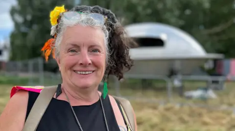 Tom Jackson/BBC Sarah Corbett is standing to the left. She has brown and grey hair in a ponytail and two flowers in her hair. She has spectacles resting on top of her head and is wearing a black vest top. In the background is a metal UFO which is part of a stage.