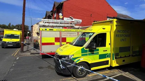 BBC/Sam Hotson An alternate view of the cordoned-off road, which shows the tape stretching around a building made from red-painted brick on the corner, and another ambulance parked down an ajacent road.
