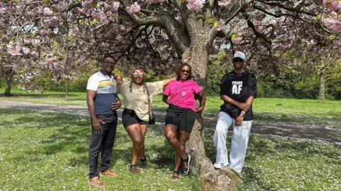 Julia Bryson/BBC A family of four - two men and two women - pose for a photo beneath a blossom tree in a park called the Stray in Harrogate .