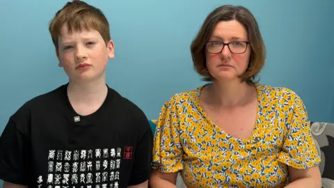 Shows a woman and young boy looking into the camera with a serious expression. The woman has short brown hair and is wearing glasses and a yellow and blue floral shirt. The boy has red hair and is wearing a black patterned shirt.