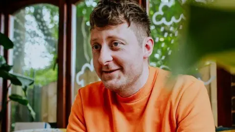 Walker Books Children's author Jarvis who has brown hair wearing an orange t-shirt sitting on a sofa in front of windows with a garden view.