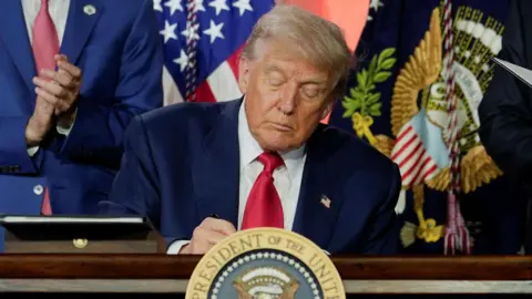 US President Donald Trump wearing a blue suit, red neck tie and white dress shirt at a desk in the White House looking down while signing executive orders.