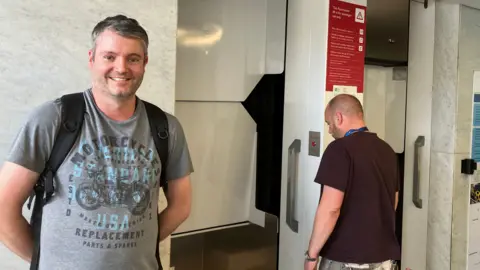 John Allred standing beside a lift in a modern interior with marble walls. He is wearing a graphic T-shirt and backpack, while another person stands near the lift controls with their back turned. Red safety signage and wall-mounted displays are visible next to the lift.