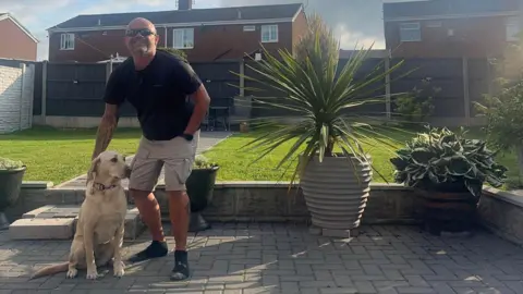 A man wearing sun glasses, a T-shirt and shorts, stands in a garden smiling with his right hand on a dog, which is sitting. Behind him is a lawn with a brown fence at the back. He is standing on a paved courtyard with flower pots on it and steps leading up to the lawn.