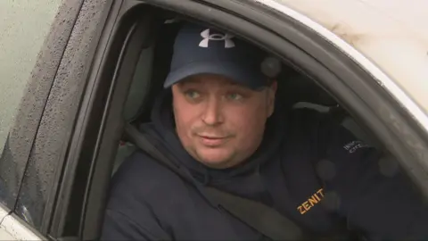 Ross Colquhoun, a contract worker at Exxon Mobil Mosmorran, sits in his car. He is wearing a navy hooded top with the Zenith logo on it and a navy blue cap. 