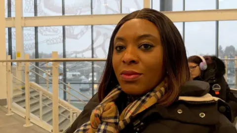 Alex Dunlop/BBC Bernadette Martins sitting in a railway station, looking at the camera, wearing a dark coat, checked scarf, with long dark hair with stairs to the left. 