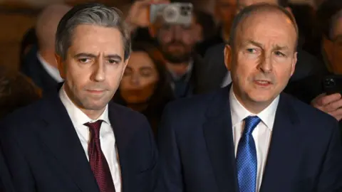 Getty Images Simon Harris, standing near Mr Martin, looks into the distance. Martin is balding and wears a dark blue suit, white shirt and royal blue tie, while Harris has short brown hair and wears a dark blue suit, white shirt and maroon tie.