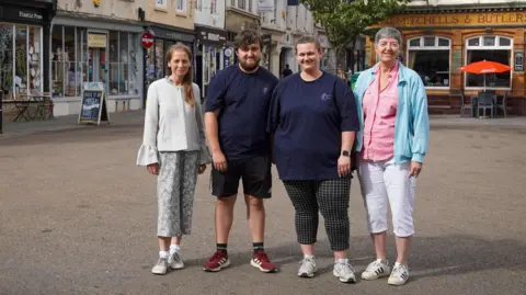 A group of four people - a man and three women - are standing in the Cornmarket in Worcester. Behind them is a row of shops and a pub.
