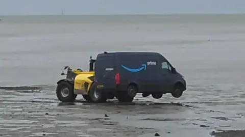 A forklift truck, or similar vehicle, is lifting a navy blue Amazon Prime branded van on mudflats. The sea is behind, and there is blue sky