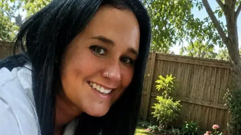 A young woman with straight dark hair, smiling. She has a nose ring and a piercing below her bottom lip. She is sitting outside in a garden on a sunny day, in front of a wooden fence and several trees.