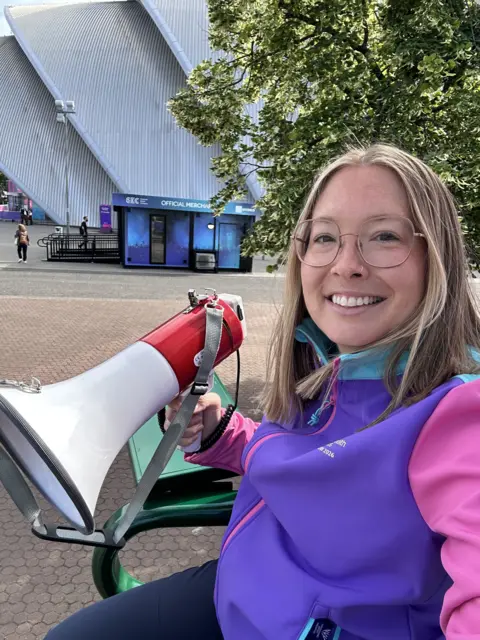 Alice McNaugher Alice McNaugher, sitting in a tall chair similar to those used by tennis umpires. She is wearing a purple and pink jacket and holding a megaphone in one hand. She is sitting outside at the SEC campus in Glasgow. 