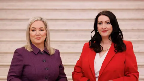 PA Media Two women stand side by side looking at the camera. The woman on the left has blonde hair and is wearing a purple jacket. The woman on the right has dark longer hair and is wearing a red jacket and white shirt. They are both stood in front of a pale marble staircase