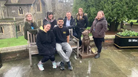 Justin Dealey/BBC Six people sitting and standing around a wooden bench with the engraved words “HOPE IS EVERYTHING” on the seat slats. Next to the bench is a bronze statue of a German shepherd dog.