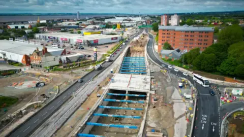 Baxter Media The road is completely dug up in the middle of the carriageway with blue steels attached to the sides of the road as they dig down to a lower level.