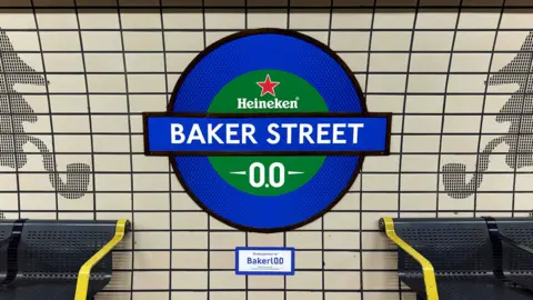 A London underground a wall sign reading “Baker Stret”, with Heineken branding inserted. The TFL sign is blue anbd green . Bakerloo is spelled with "0.0" instead of "oo" at the end
