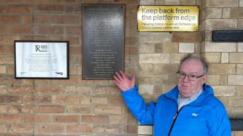 A man wearing glasses and wearing a blue coat, grey jumper and white and blue checked shirt stands in front of a plaque on a station platform. The plaque reads In memory of Henry West, it is hung on the light brown brick wall. A prominent yellow sign in the top right hand corner reads "Keep back from the platform edge. Passing trains cause air turbulence. Stand behind the yellow line"