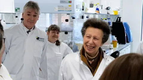 Princess Anne wearing a white lab coat at the Quadram Institute in Norwich. She is looking off from the camera and is smiling. Two other people in white lab coats are looking on behind her.