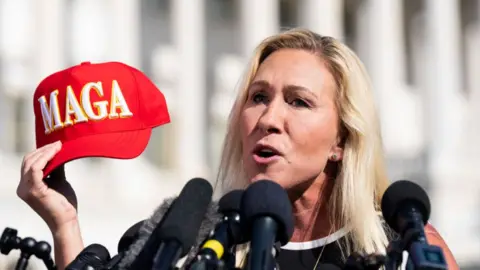 Image shows Marjorie Taylor Greene holding a MAGA hat