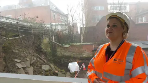 Karen Shore, Cheshire West and Chester Council's deputy leader, is photographed in front of the city walls. She is wearing an orange hi-vis jacket and white hard hat.