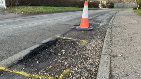 A generic image of a pothole damaged road with yellow paint sprayed around the hole and an orange traffic cone warning drivers of the damaged road