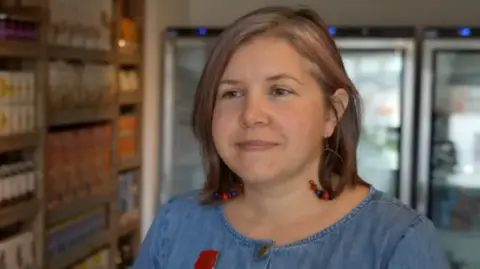 A woman with a bob wearing a denim dress, hooped earrings standing in a blurred background shop.