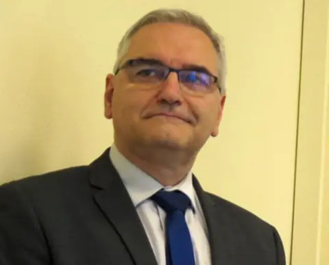 Grand Est Region & Bas-Rhin Prefect A man in a dark suit jacket, pale shirt and blue tie with short grey hair. He is wearing glasses. There is a plain cream-coloured wall behind him.